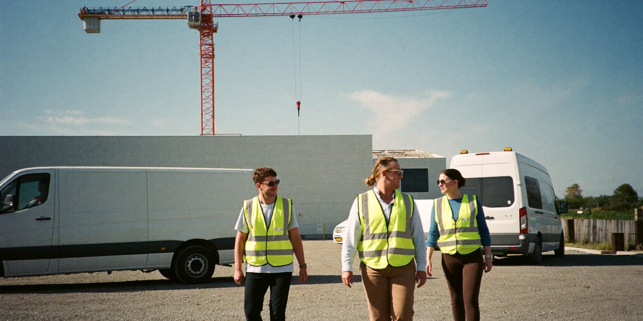 Techniker in gelben Warnwesten gehen über einen Schotterplatz von weißen Transportern zu einem Rechenzentrum im Bau