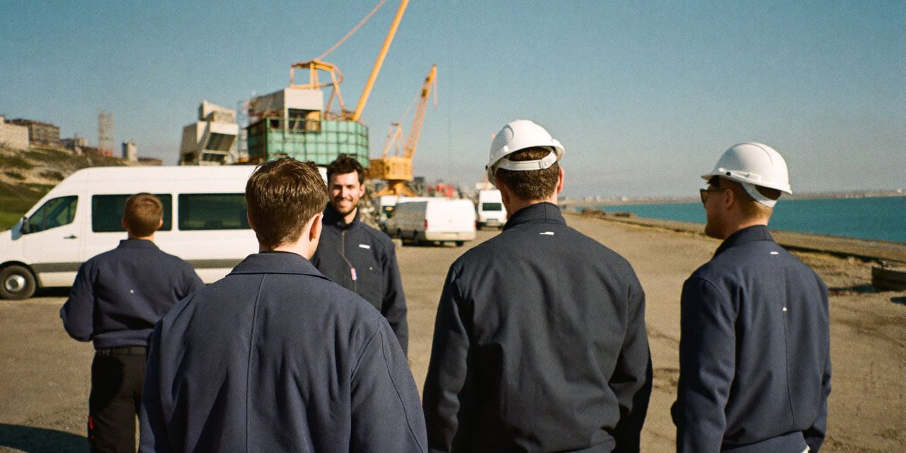 Elektriker in dunkelblauen Arbeitsjacken im Gespräch auf einer Baustelle im Hafen, mit Kränen und weißen Transportern
