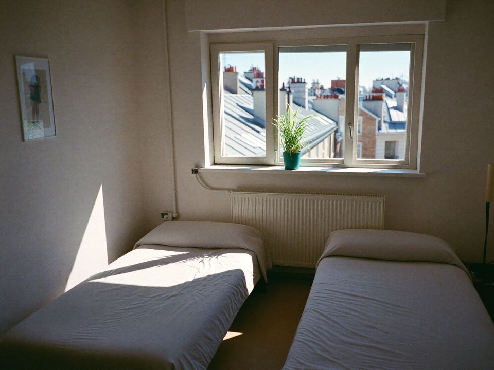 Helles Zweibettzimmer mit zwei gemachten Betten und einem Fenster mit Blick über die Dächer